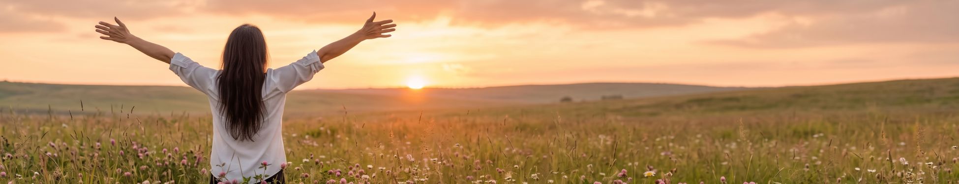 ujer de espaldas con los brazos abiertos en un campo de flores al atardecer, en una escena de calma, libertad y bienestar emocional.