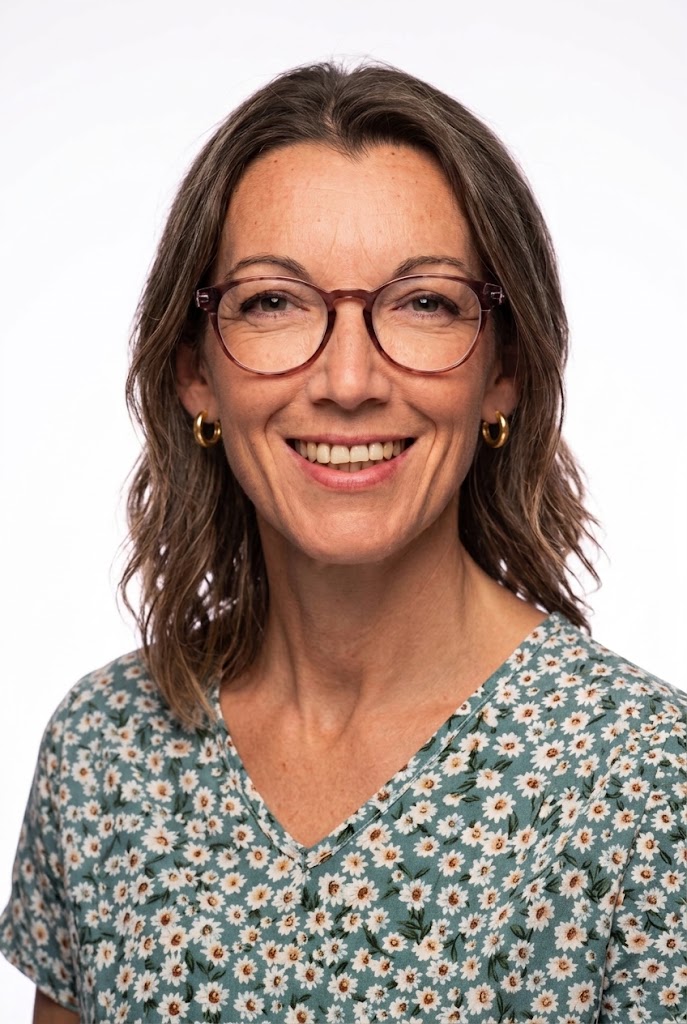 Retrato de una mujer sonriente con gafas y pendientes, de pelo castaño ondulado hasta los hombros, vestida con una camisa de flores verdes  sobre fondo blanco.