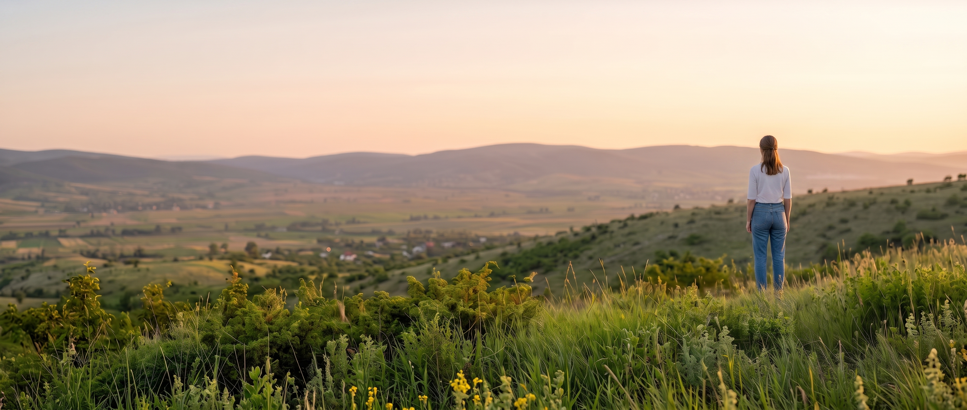 Mujer de espaldas contemplando un amplio paisaje de campos y montañas al atardecer.